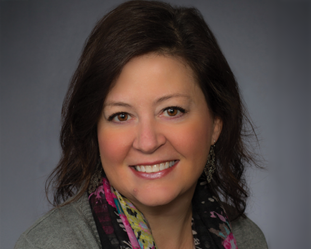 Professional headshot of a woman with dark hair wearing a gray blazer and colorful scarf